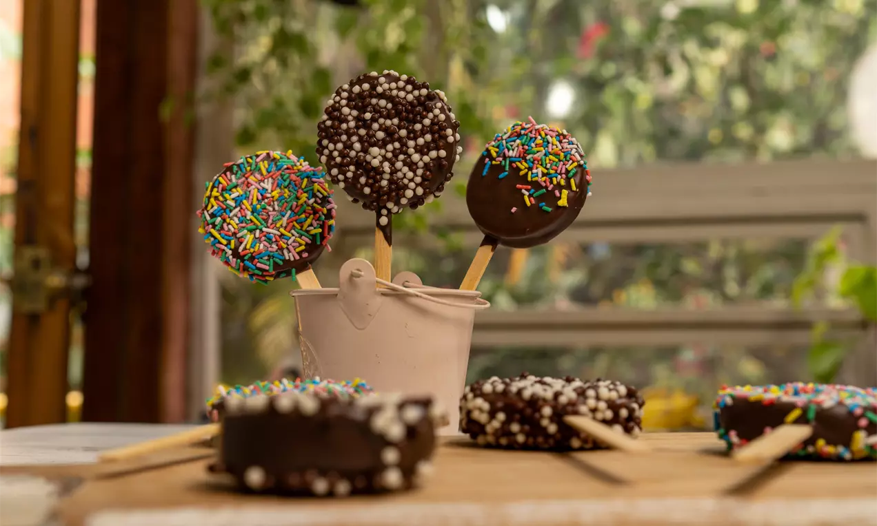 A imagem mostra pirulitos de biscoito cobertos com chocolate, decorados com confeitos coloridos e bolinhas crocantes.