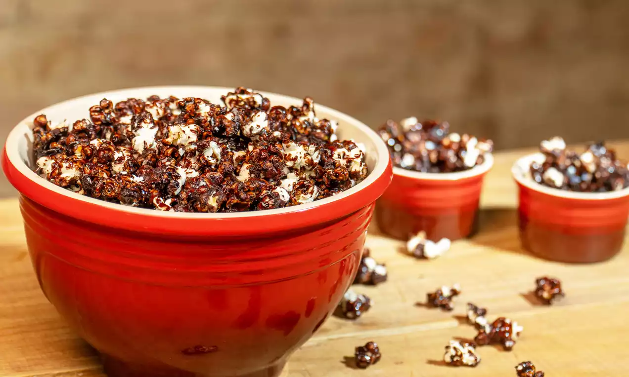 Pipocas brancas envoltas em camada irregular de chocolate escuro brilhante dentro de tigela vermelha com algumas espalhadas na mesa de madeira clara
