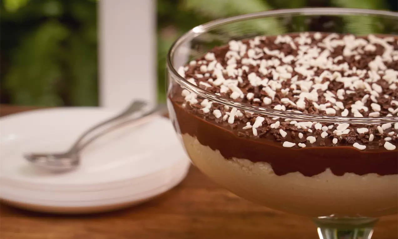 Creme bege suave em taça de vidro, coberto por camada de chocolate brilhante e finalizado com flocos brancos e granulados escuros