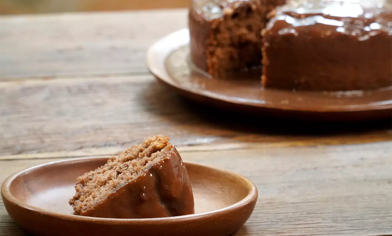 Fatia de bolo marrom macio com cobertura brilhante de brigadeiro de avelã em prato de madeira diante do bolo inteiro sobre mesa rústica
