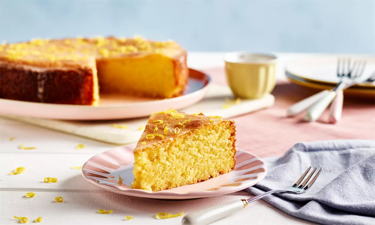 Fatia de bolo fofo de limão amarelo com cobertura brilhante e raspas de limão em prato rosa, sobre mesa com guardanapo lilás e talheres