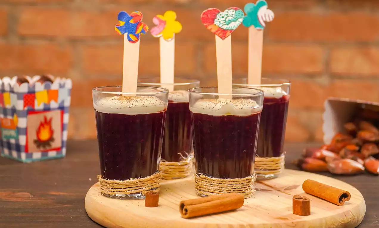 Copos transparentes com sagu roxo brilhante decorados com palitos coloridos e borda enlaçada sobre tábua de madeira junto a canelas