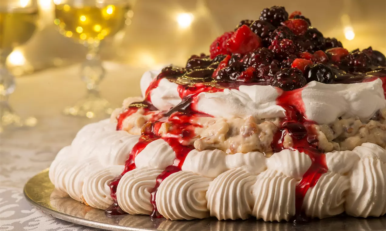 Merengue branco decorado com creme e frutos vermelhos brilhantes sobre calda rubi, dispostos em um prato prateado iluminado ao fundo