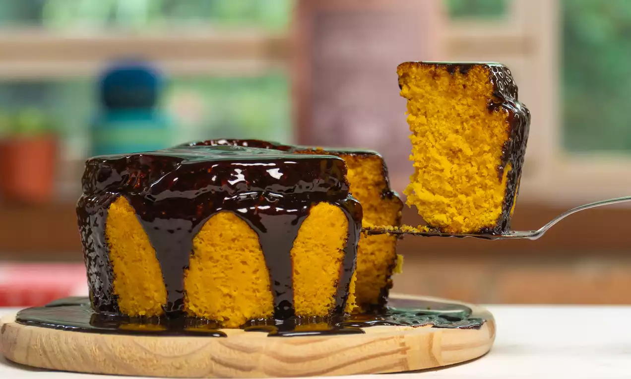 Bolo de cenoura fofo coberto com glacê de chocolate brilhante, fatia servida em prato branco com raspas de chocolate e cenoura ralada
