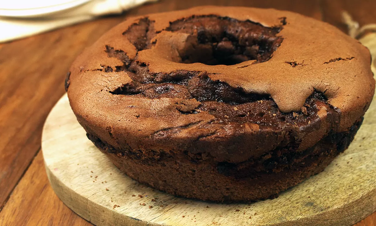 Bolo redondo com textura macia, mescla marrom e chocolate em tons claros e escuros, servido sobre tábua de madeira