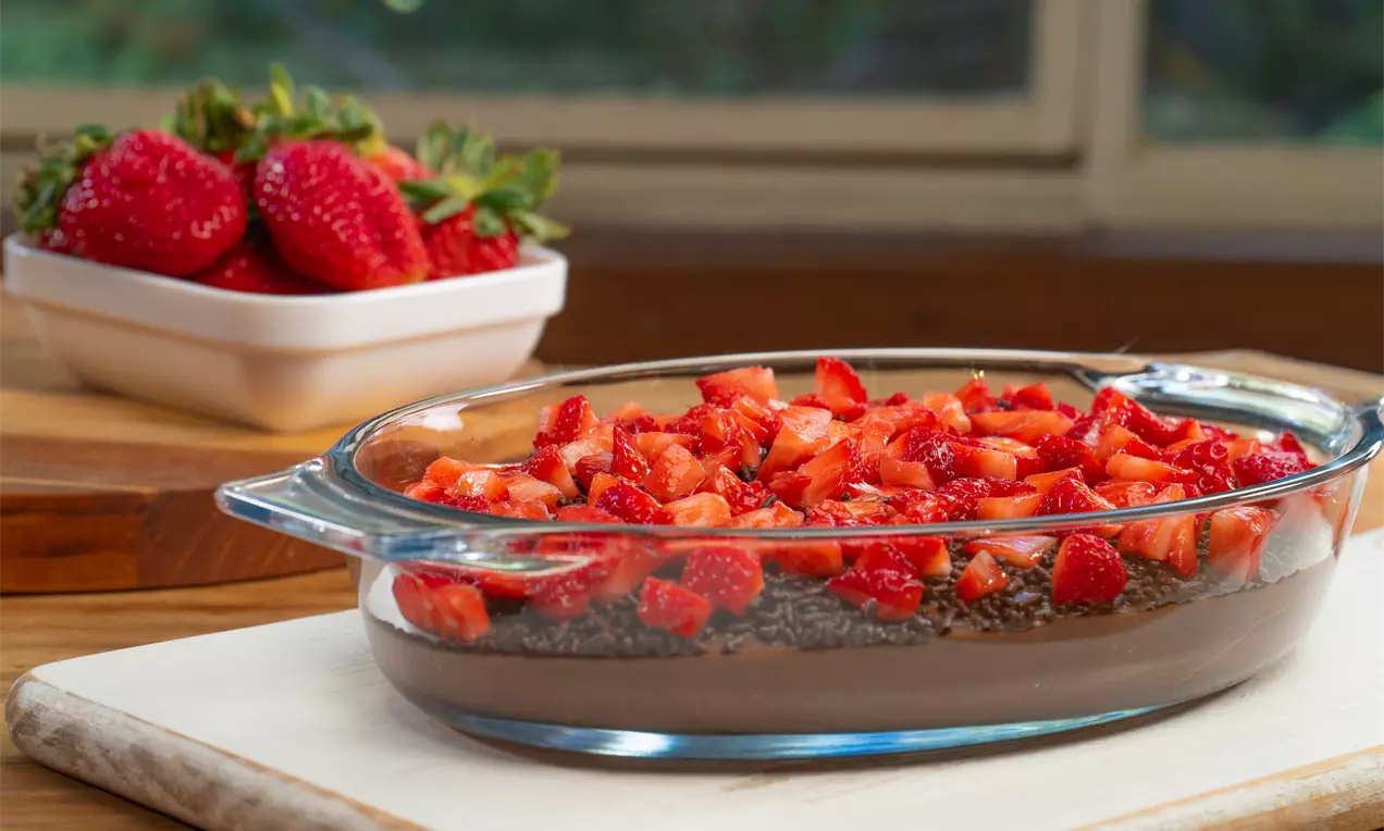 Brigadeirão cremoso de chocolate escuro em travessa de vidro, coberto com granulado brilhante e pedaços frescos de morango vermelho