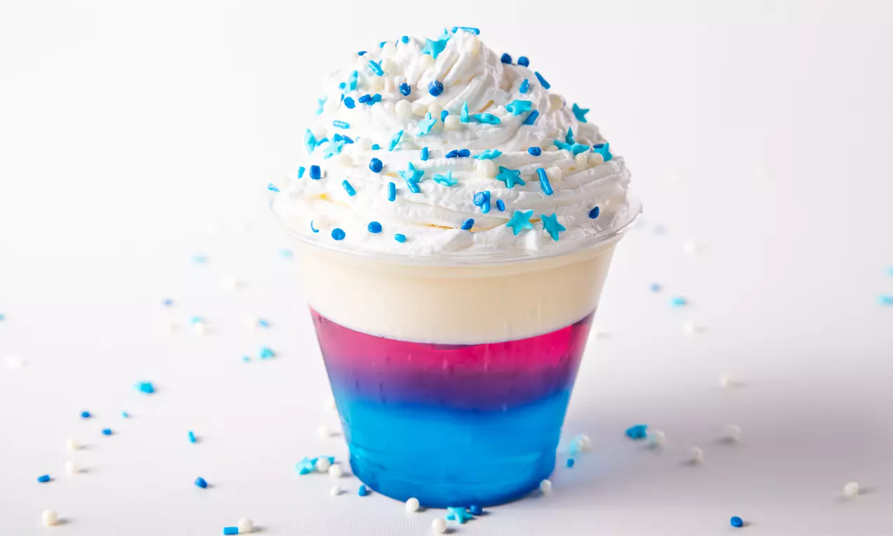 Gelatina em camadas azul e rosa vibrantes com creme branco aerado no topo, coberto por confeitos azuis e brancos em um copo transparente