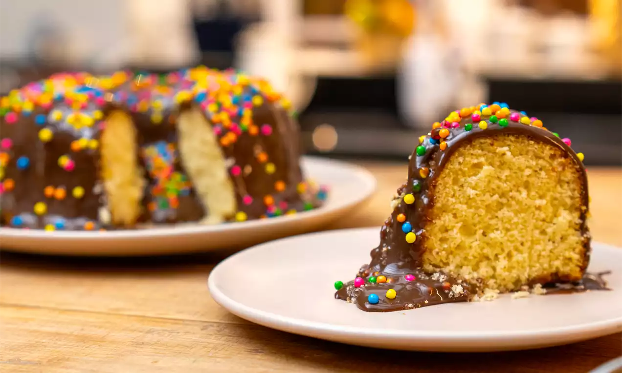 Bolo em forma de anel com massa amarela fofa, cobertura lisa de chocolate e confeitos coloridos, servido em pratos brancos sobre mesa de madeira