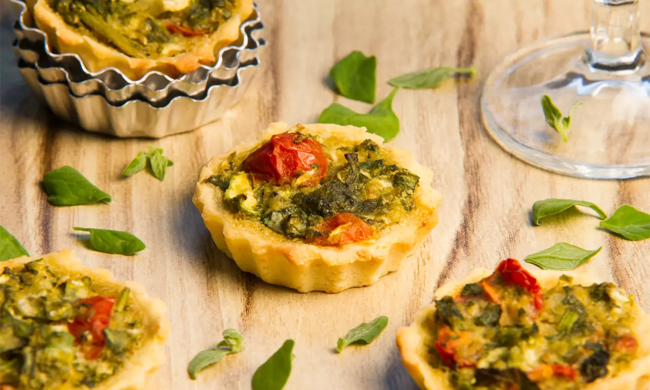 Mini quiches sem glúten com bordas douradas e crocantes, recheio verde com espinafre e pedaços de tomate sobre mesa de madeira clara