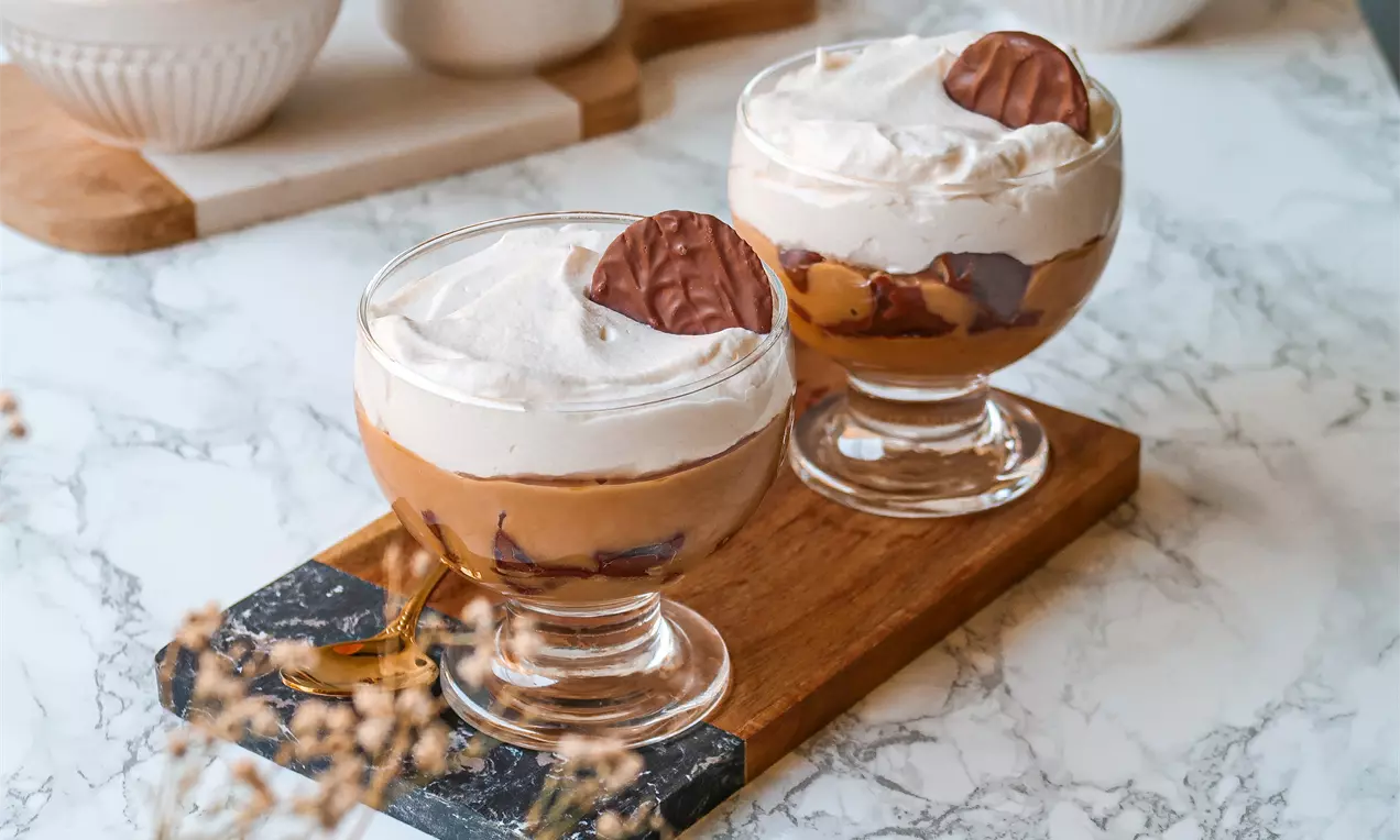 Taças de vidro com camadas de creme caramelo bege, chocolate escuro e chantilly branco, decoradas com biscoito redondo em base de madeira