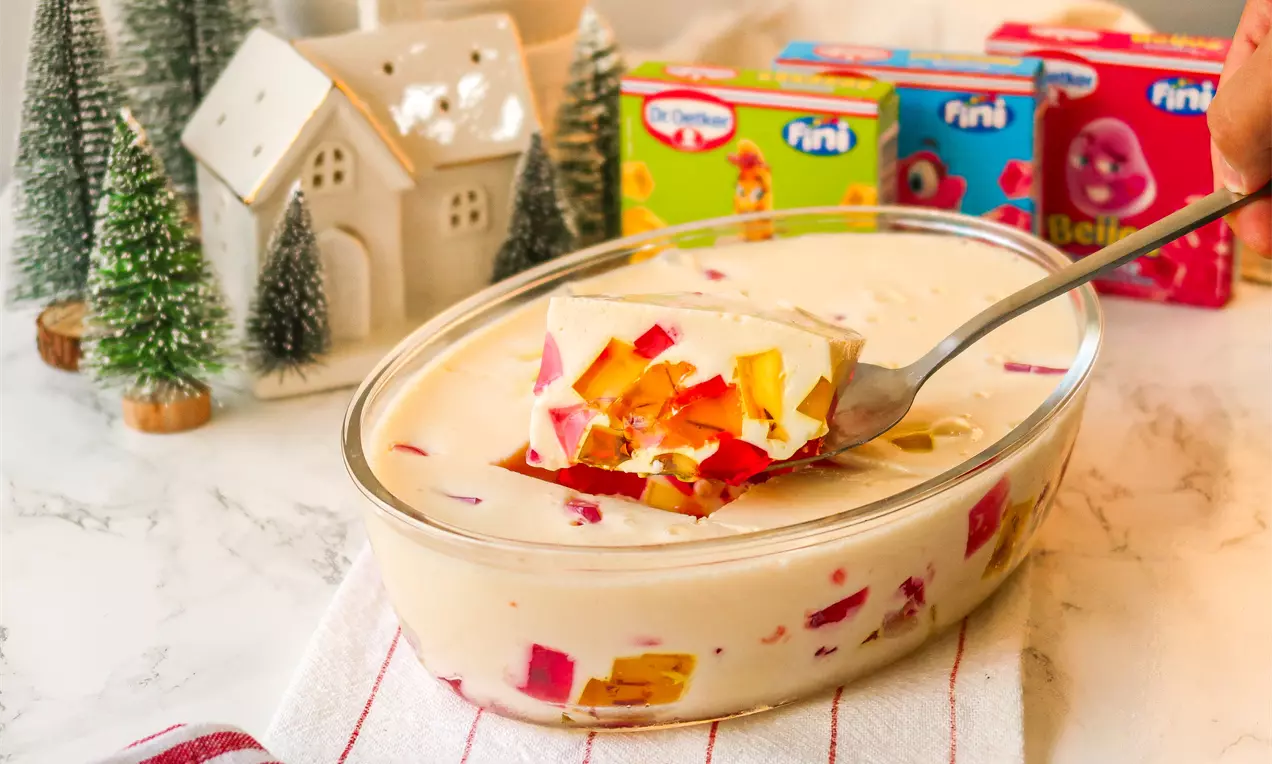 Gelatina colorida em cubos vibrantes de amarelo, laranja e vermelho misturada a um creme branco em travessa oval sobre toalha listrada