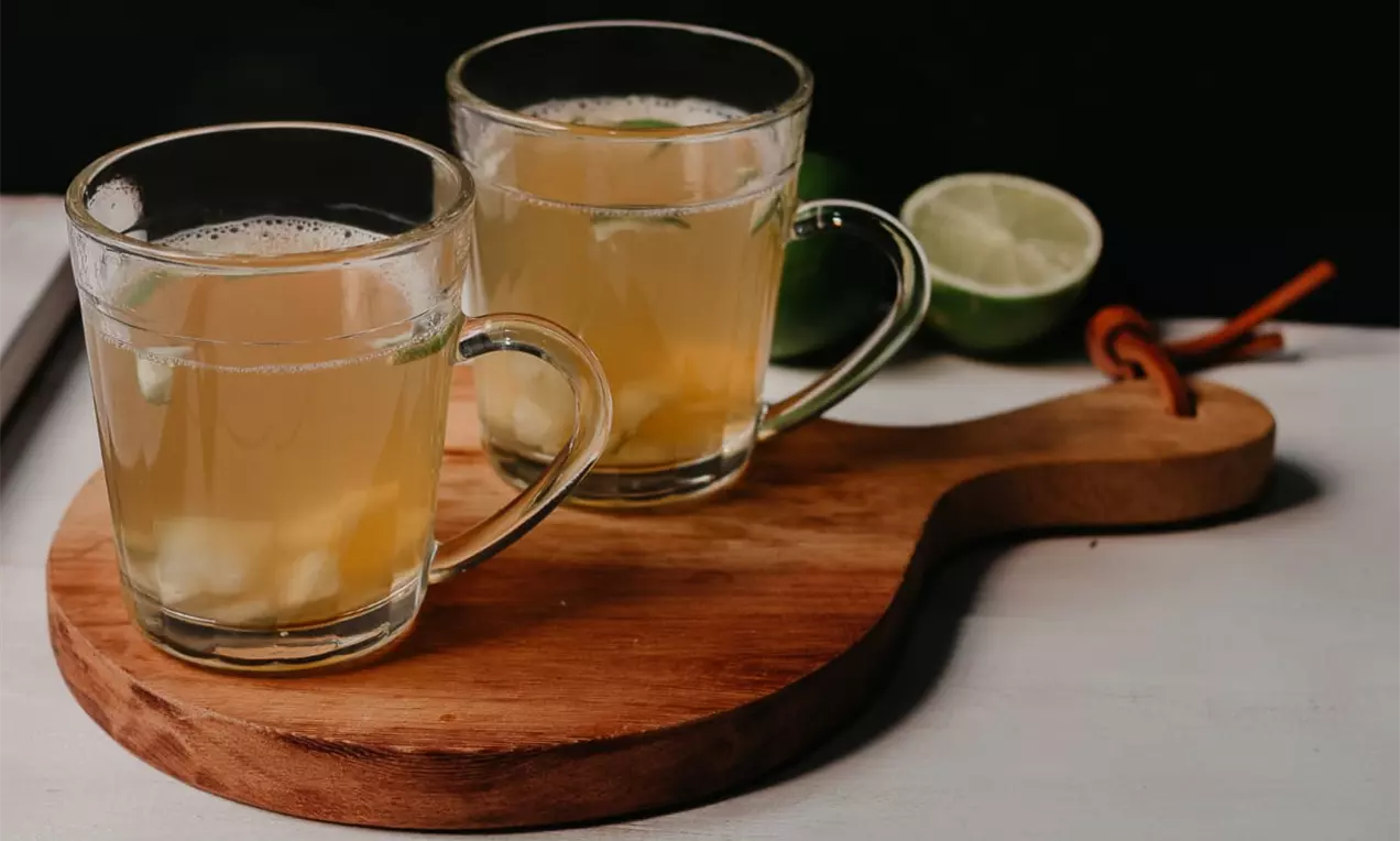Chá amarelo claro com pedaços de abacaxi e folhas verdes em xícaras de vidro sobre tábua de madeira acompanhado de limão cortado