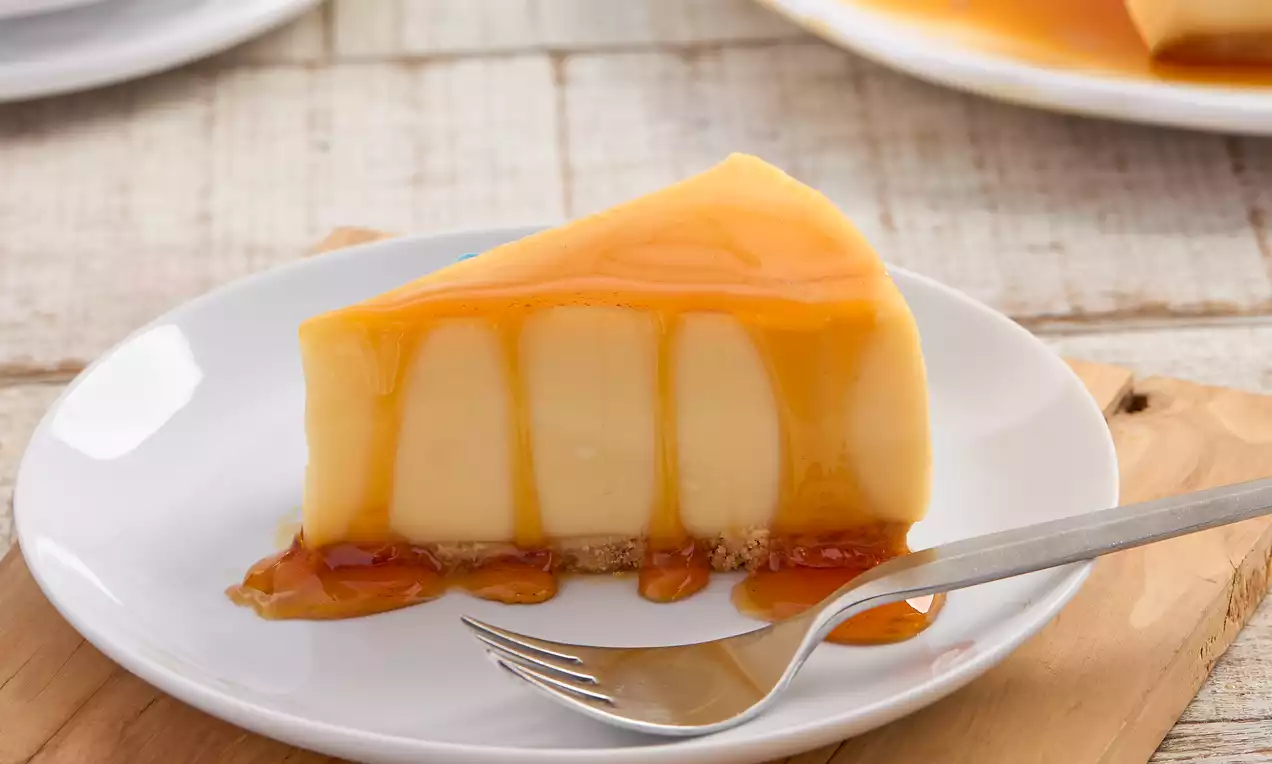 Fatia de torta cremosa alaranjada com cobertura de calda caramelada brilhante e base de bolacha sobre prato branco com garfo prata