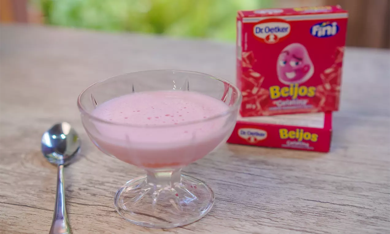 Sobremesa cremosa de gelatina rosa clara com textura leve e lisa servida em uma taça de vidro transparente sobre mesa de madeira clara