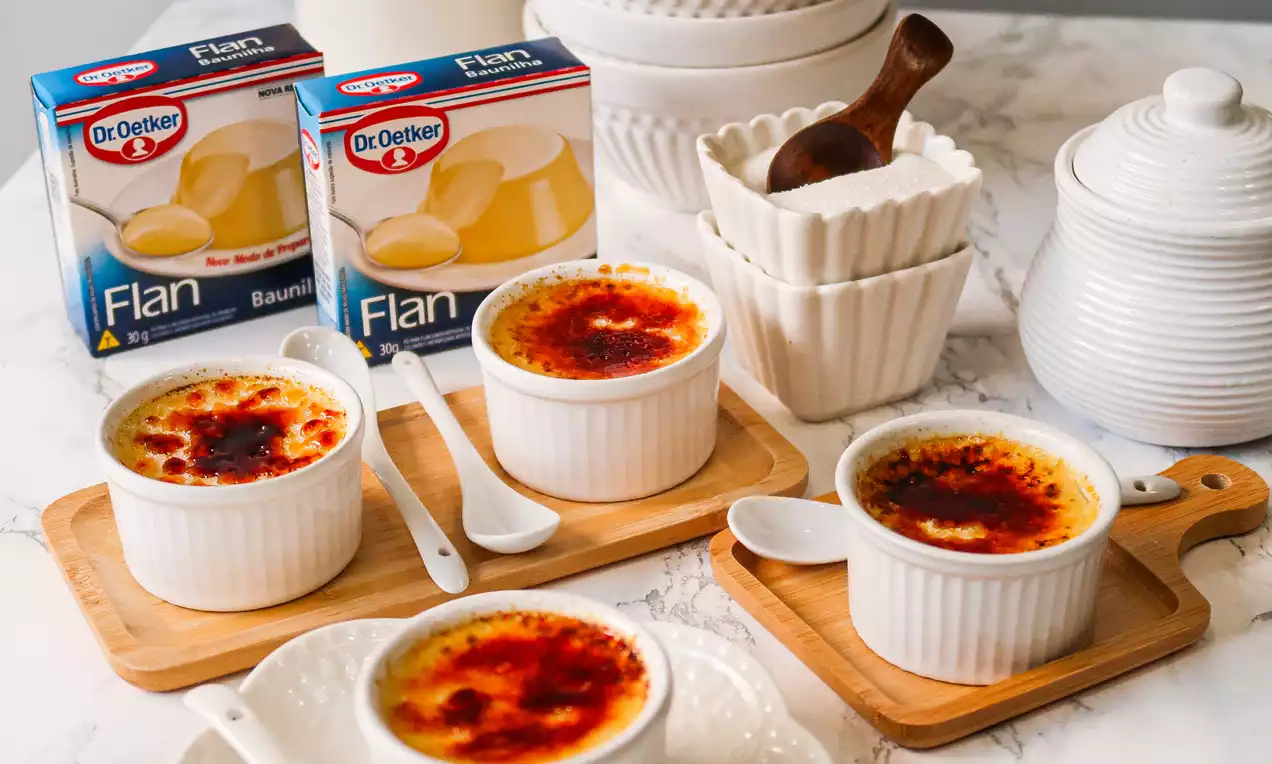 Flans dourados com crosta caramelizada irregular em tigelas brancas estriadas, dispostos sobre tábuas de madeira com colheres ao lado