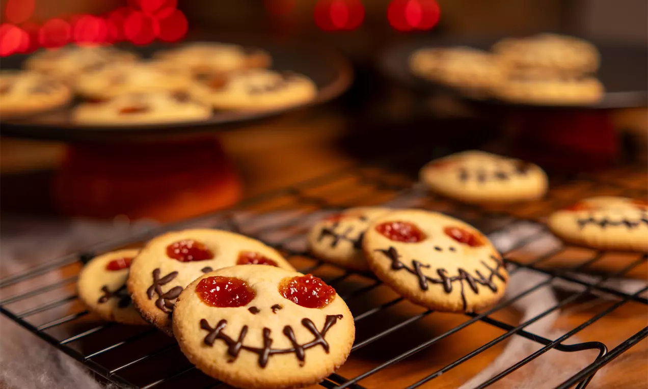Biscoitos de Halloween em forma de rostinhos assustadores, com olhos de geleia vermelha e bocas desenhadas com chocolate, sobre uma grade de resfriamento iluminada por luzes vermelhas.