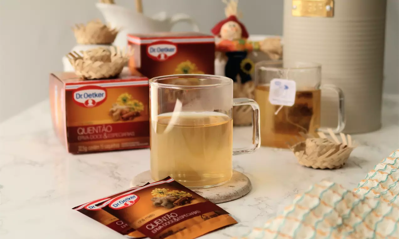 Chá dourado translúcido servido em caneca de vidro sobre base de mármore clara, acompanhado por sachês e decoração rústica em fundo neutro