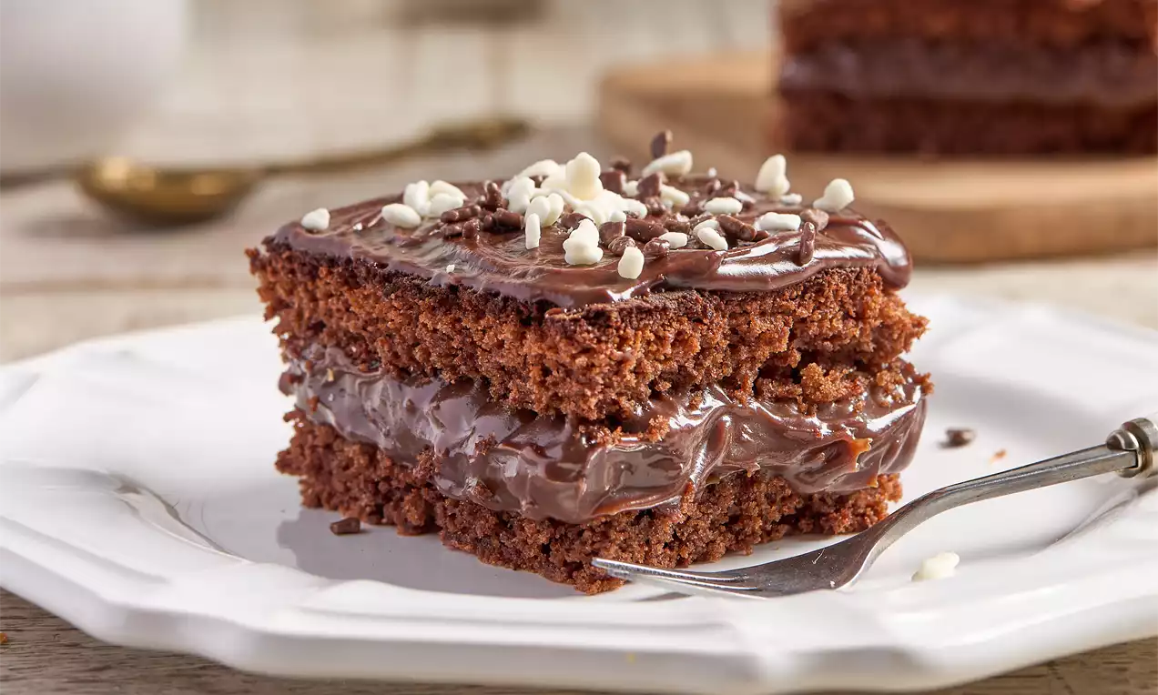 Fatia quadrada de bolo de brigadeiro com camadas de massa marrom aerada, recheio cremoso e cobertura brilhosa, decorada com flocos brancos e escuros sobre prato branco