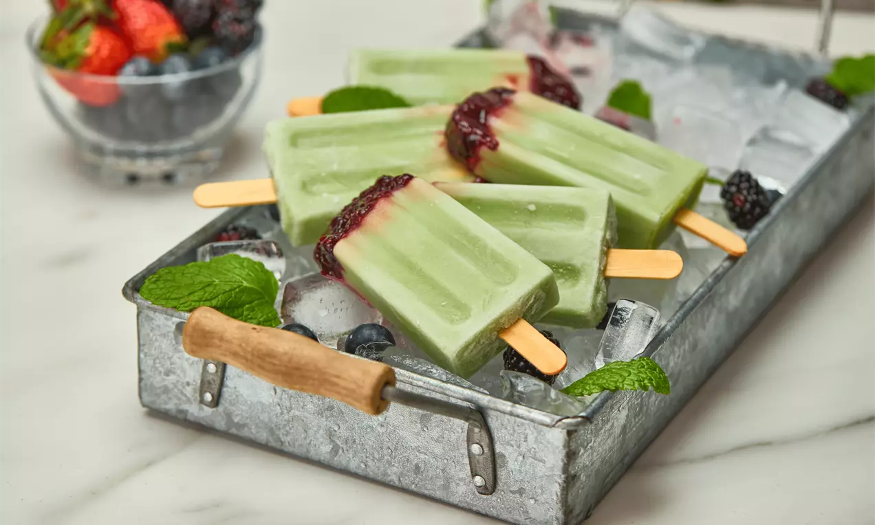 Picolés retangulares verde-claro cremosos com geléia de frutas vermelhas no topo, dispostos sobre gelo em bandeja metálica com folhas de hortelã