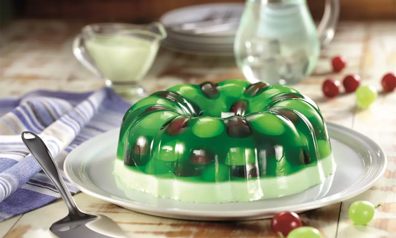 Gelatina verde translúcida em formato circular, com uvas vermelhas e verdes suspensas, sobre camada cremosa e prato branco ajustado ao fundo