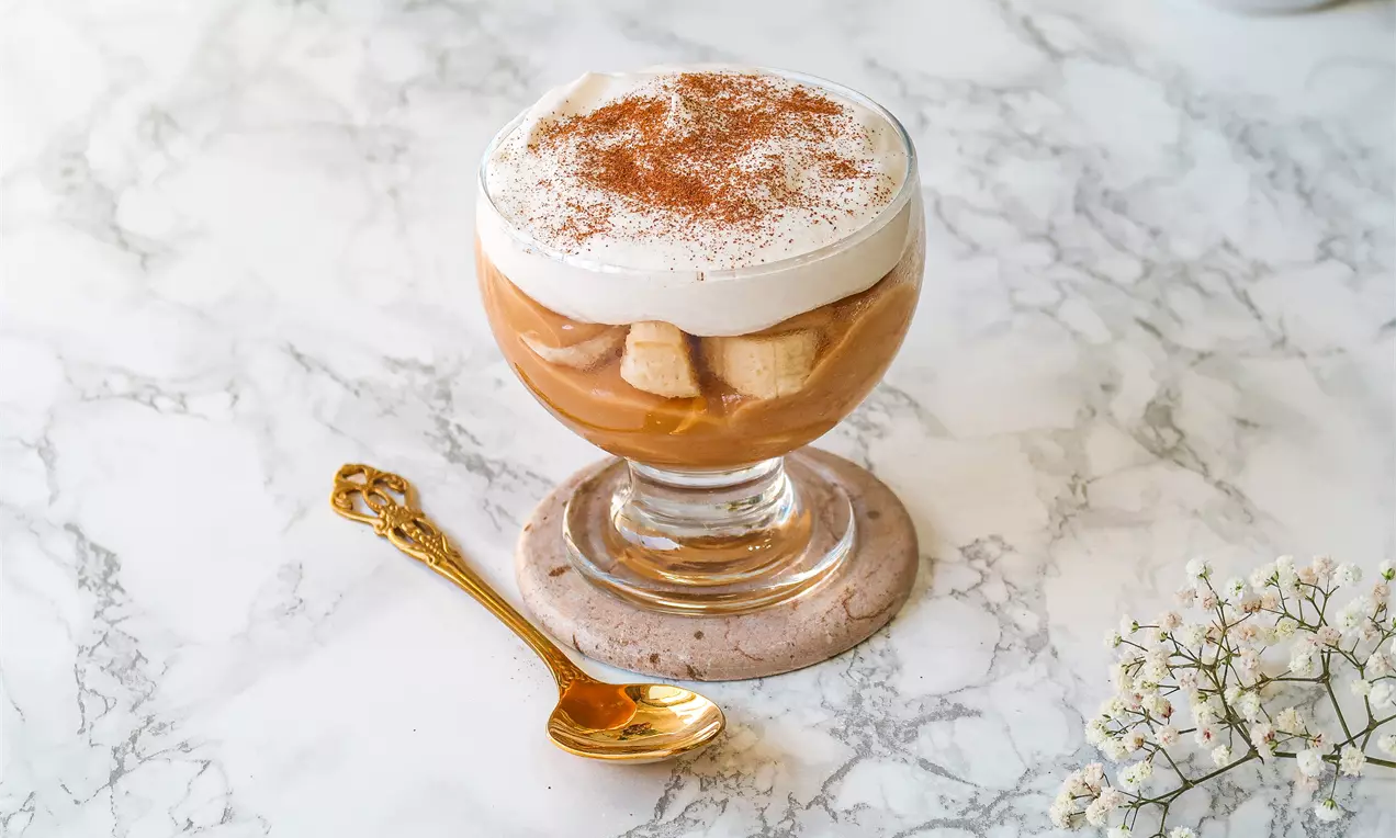 Creme caramelizado marrom com fatias de banana cobertas por chantilly branco polvilhado com canela em uma taça sobre base de mármore