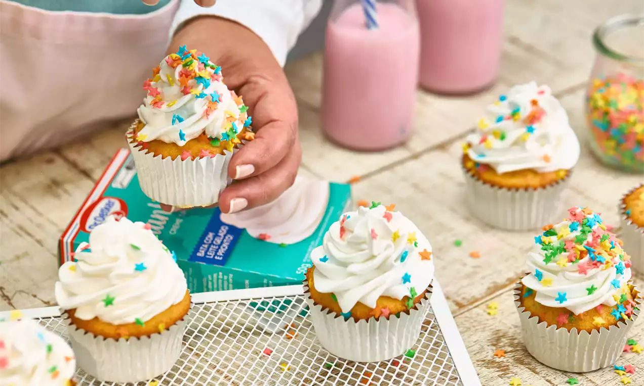 Cupcake dourado de topo arredondado coberto com creme branco em espirais e confeitos coloridos em forma de estrelas sobre mesa clara