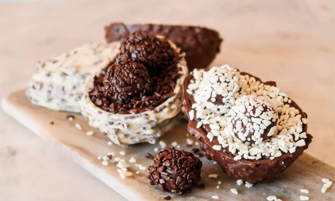Metade de ovo de chocolate ao leite com textura crocante, recheado com trufas cobertas de confeitos brancos e marrons em uma tábua de madeira clara