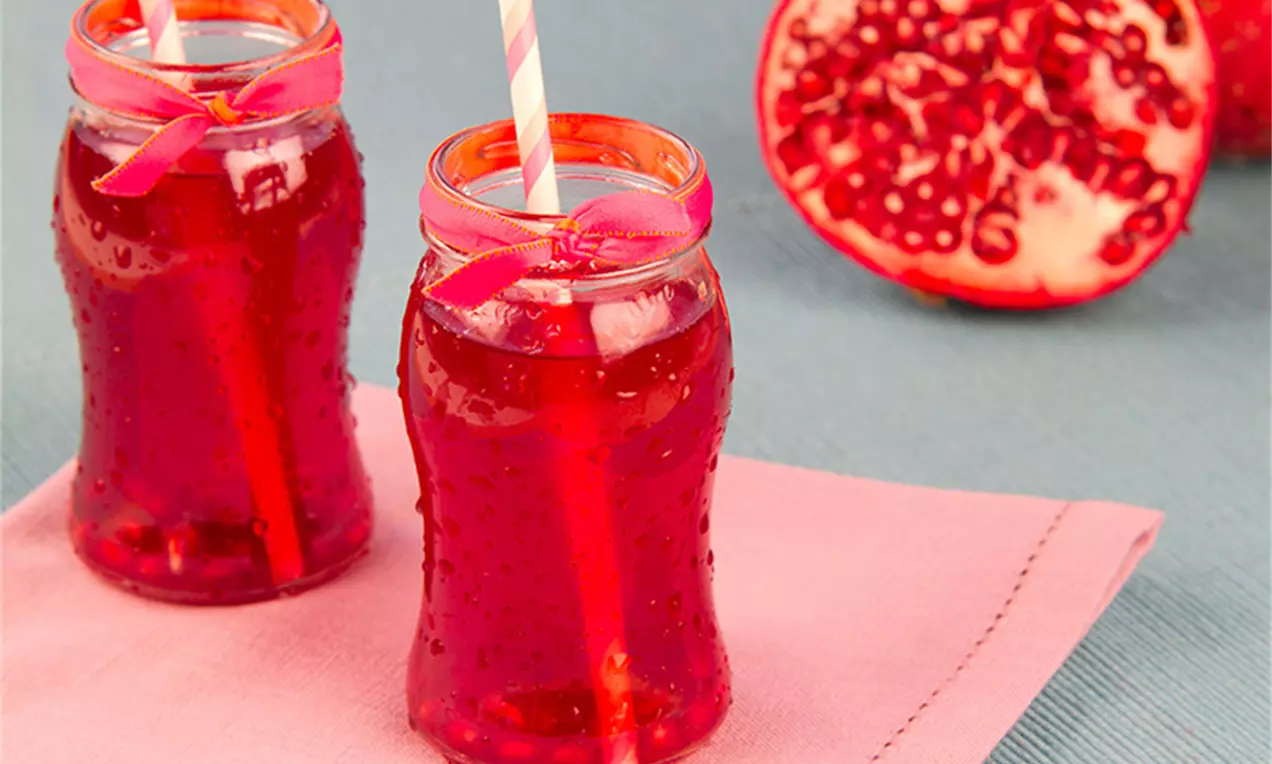Dois frascos de vidro ondulados com líquido vermelho translúcido, decorados com fitas rosa, canudos listrados e romãs abertas ao fundo