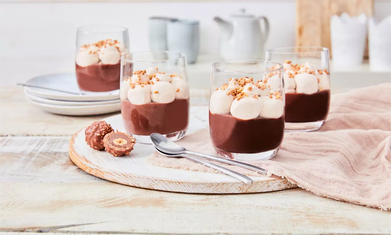 Pudim cremoso de chocolate escuro em copos transparentes decorado com camadas de chantilly branco e avelãs moídas sobre mesa de madeira clara