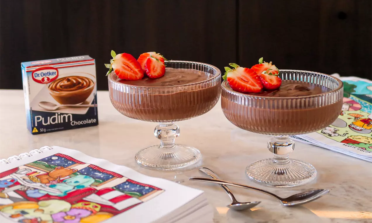 Dois potes de cristal com pudim de chocolate cremoso marrom, decorados com metades de morangos vermelhos e folhas verdes, sobre uma mesa clara