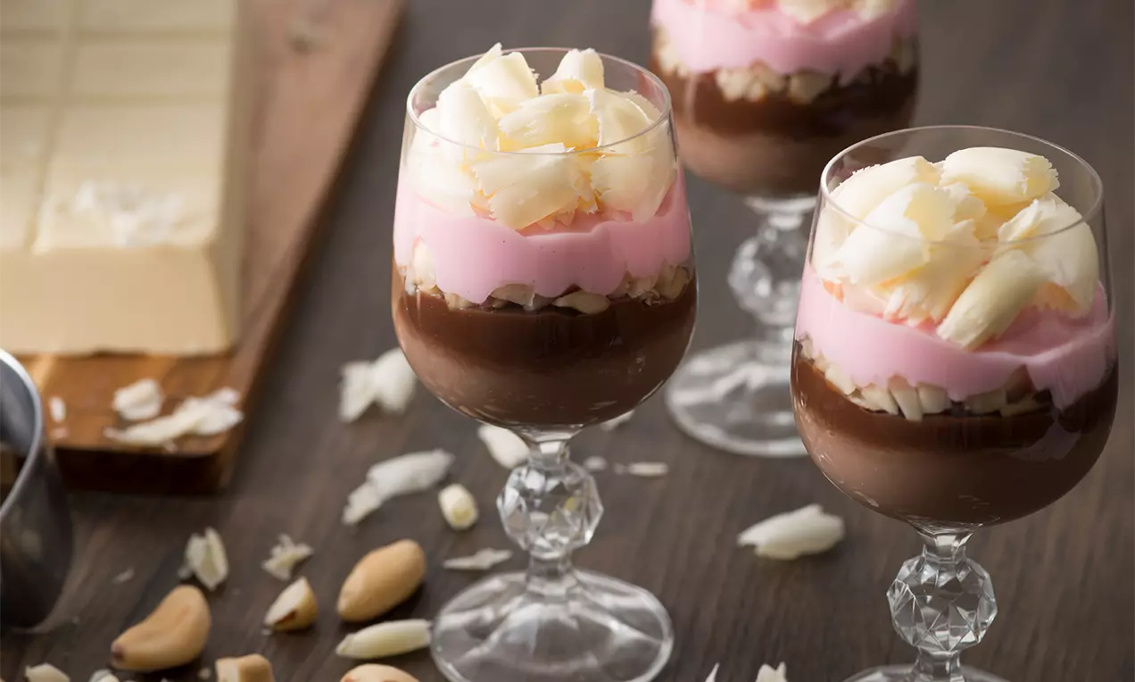 Taça de vidro transparente com camadas de creme rosa pálido, chocolate marrom e raspas de chocolate branco sobre base clássica