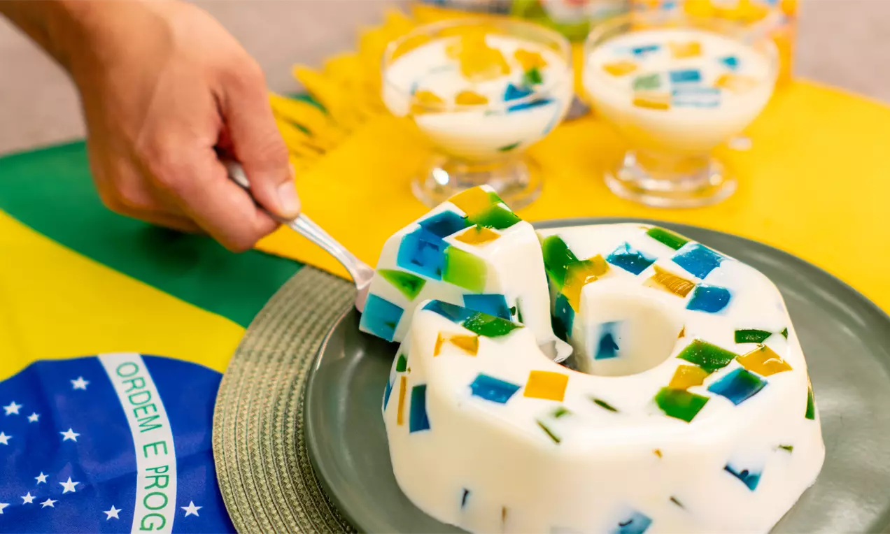 Gelatina em forma de anel com base branca cremosa e cubos coloridos verde, amarelo e azul, sobre prato verde ao lado de taças claras