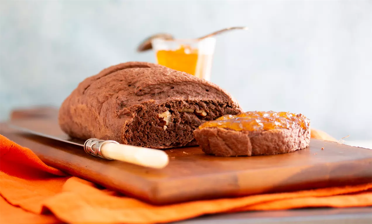 Pão marrom-escuro de cacau com textura macia, fatia aberta com geleia de laranja brilhante, faca de cabo claro em tábua de madeira