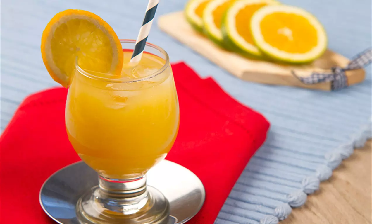 Drink amarelo vibrante em taça baixa com canudo listrado azul e branco, adornada com rodela de laranja, sobre toalha vermelha em base prateada