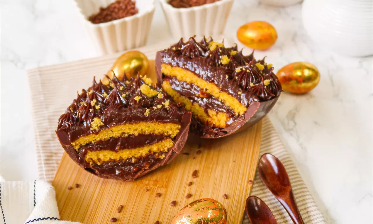 Metade de ovo de páscoa com casca de chocolate contendo camadas alternadas de bolo de cenoura úmido e creme de chocolate brilhoso sobre tábua de madeira