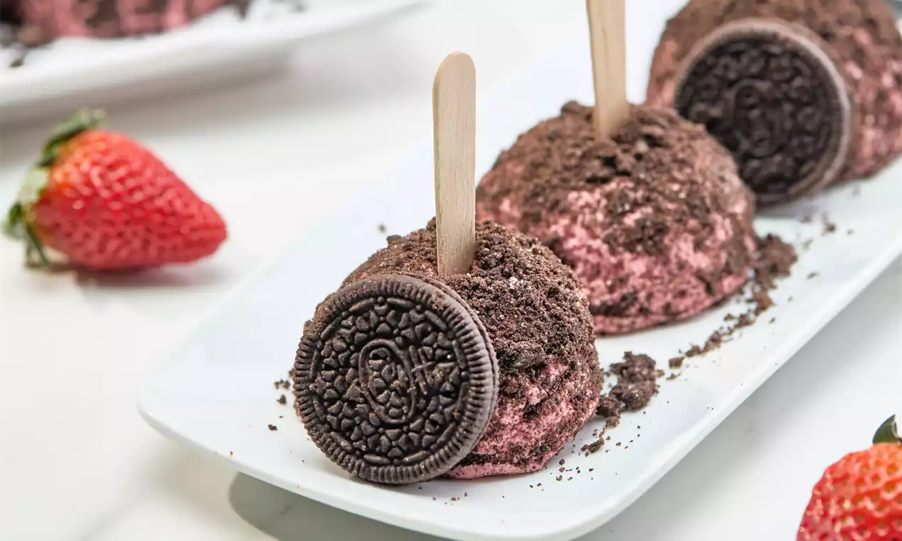 Trufas de sorvete rosa com textura cremosa, cobertas por farelos escuros, decoradas com biscoito e espetos de madeira em prato branco