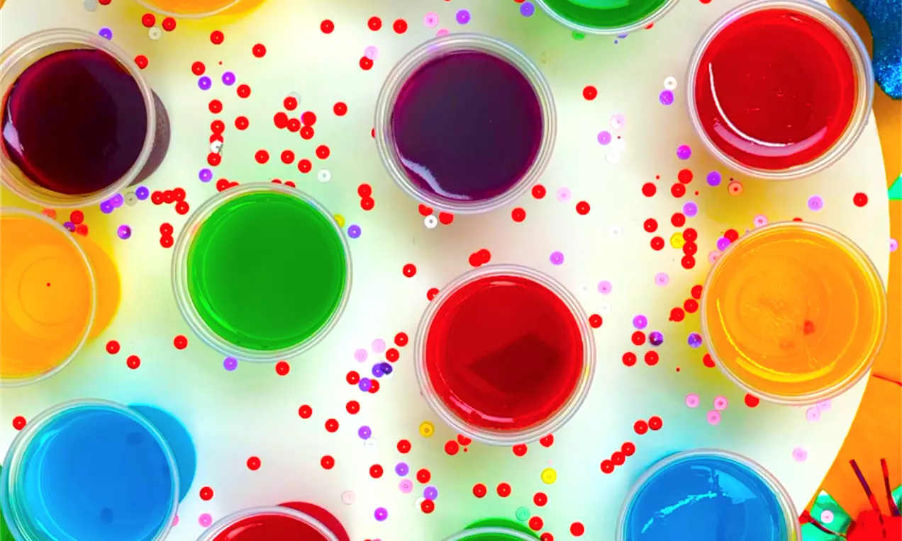 Copinhos translúcidos com geleias brilhantes em cores vibrantes, dispostos em círculo sobre prato branco decorado com confetes coloridos
