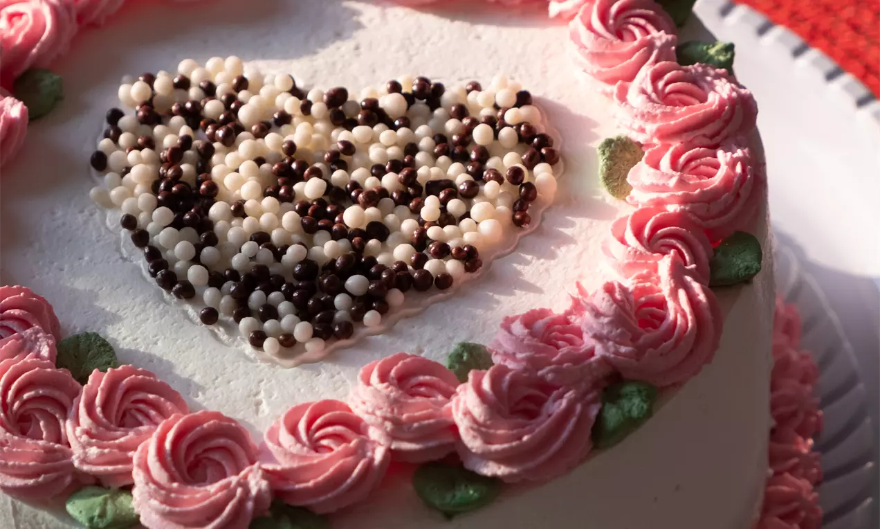 Bolo coberto com creme branco liso, decorado com rosas de chantilly rosa e folhas verdes, com um coração de confeitos brancos e escuros no topo