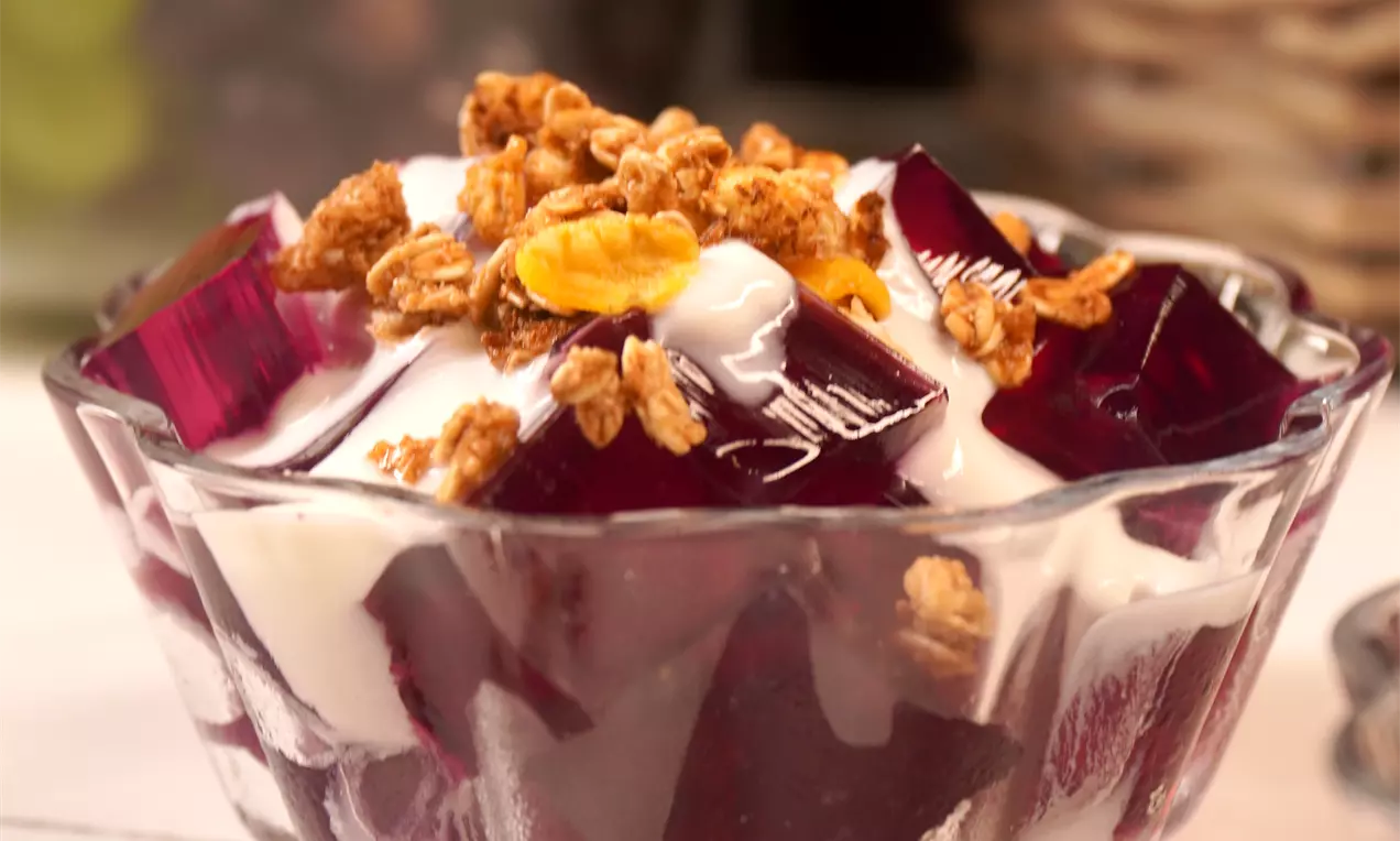Cubos translúcidos de gelatina roxa com brilho suave, entre camadas de creme branco e granola dourada em uma taça de vidro lapidado