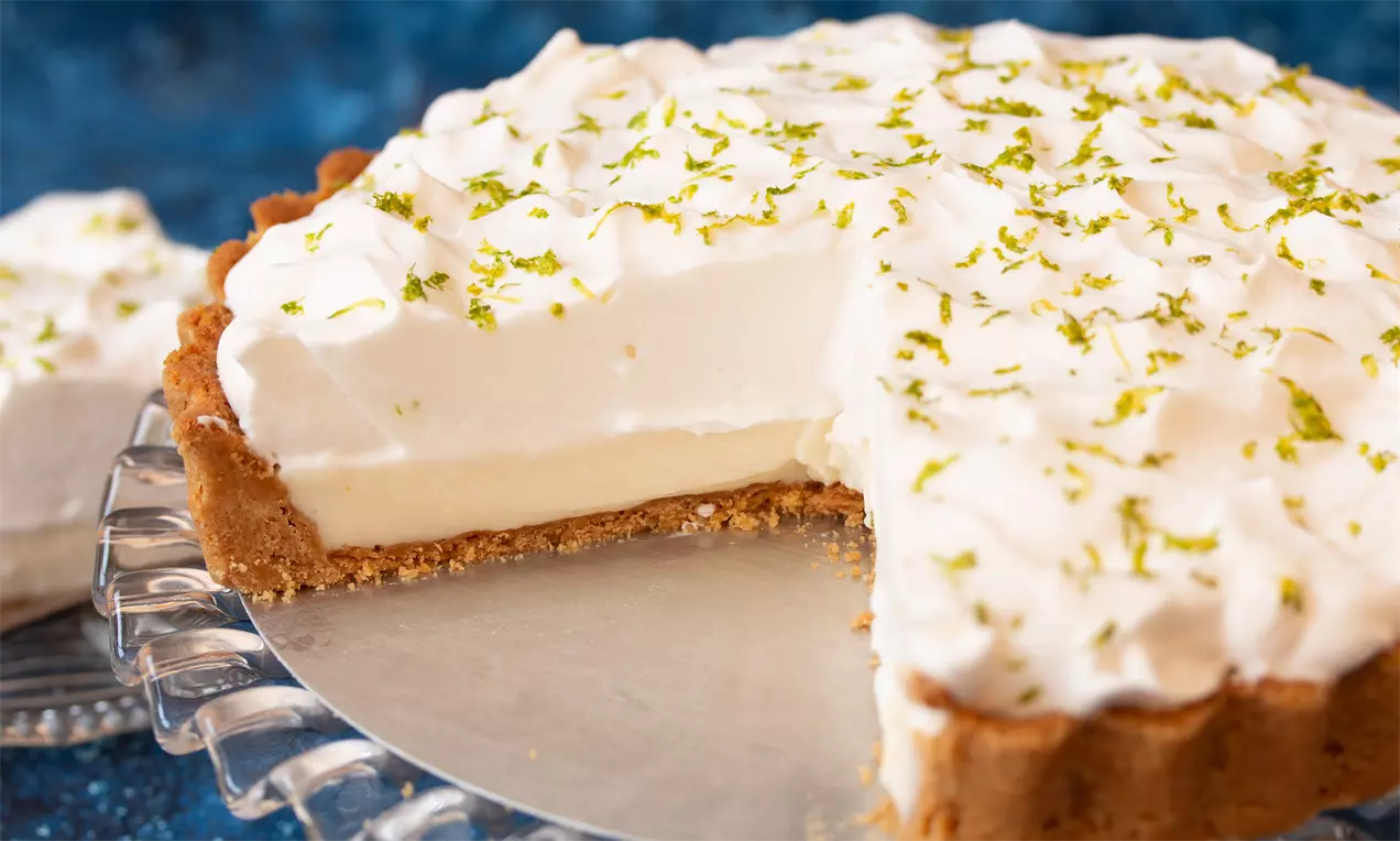 Torta de limão com massa dourada, recheio cremoso amarelo-pálido, cobertura de merengue branco levemente dourado e raspas de limão verde