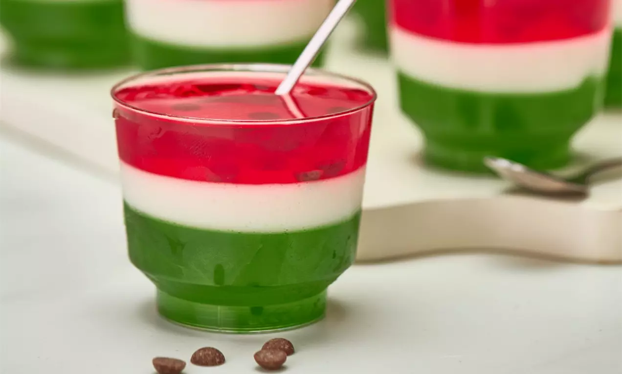 Gelatina em camadas verdes translúcidas, branca cremosa e vermelha brilhante em um copo transparente com colher prateada e gotas de chocolate