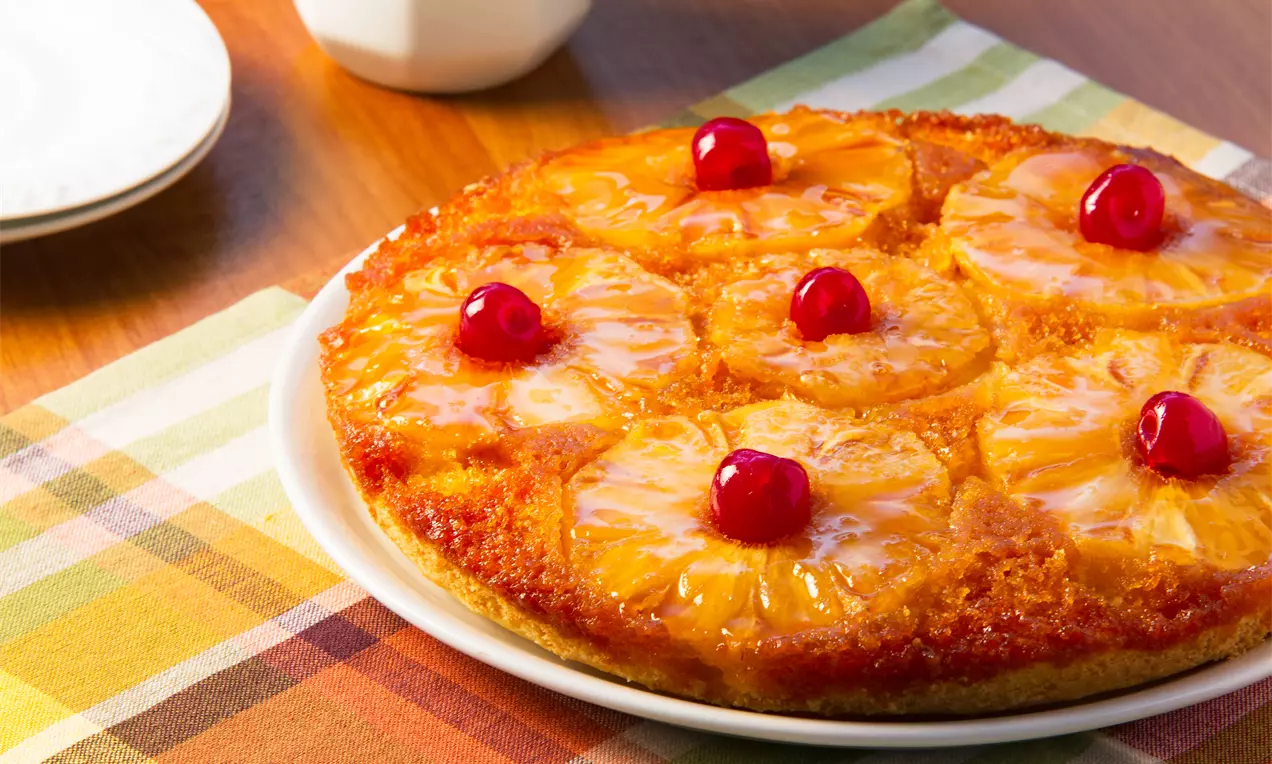 Bolo dourado com fatias caramelizadas de abacaxi em formato de anéis, decorado com cerejas vibrantes sobre prato branco em toalha colorida