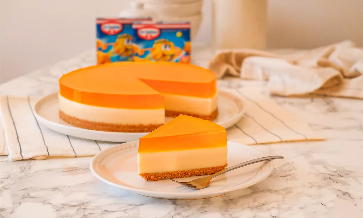 Torta circular em camadas de base marrom crocante, creme branco suave e cobertura laranja brilhante, servida em prato branco sobre mesa marmorizada