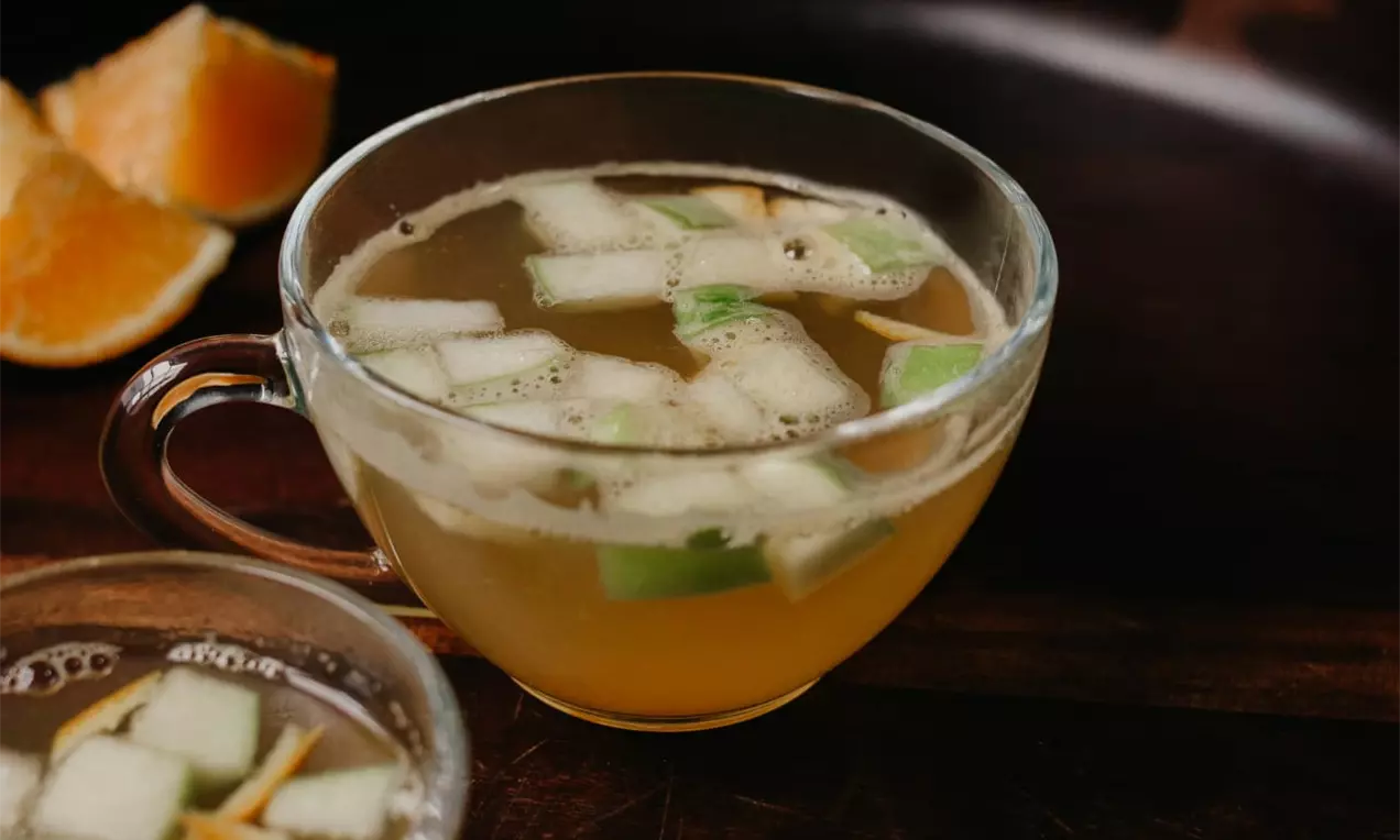 Chá dourado translúcido em xícara de vidro com pedaços de maçã verde e cascas de laranja, acompanhado por gomos de laranja ao lado