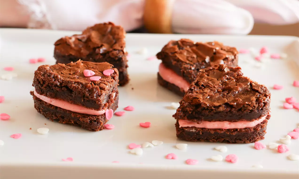 Brownies em forma de coração com textura densa e crocante, recheados com creme rosa e decorados com confeitos rosa e brancos sobre prato branco