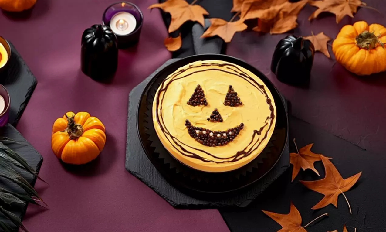 Torta de maracujá decorada com rosto de abóbora de Halloween, cercada por pequenas abóboras, velas e folhas secas sobre uma mesa escura com clima de halloween.