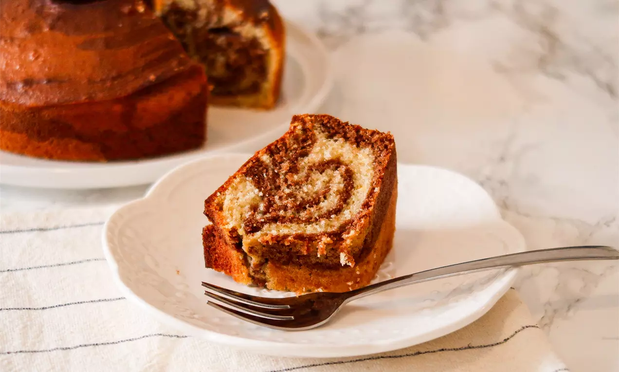 Fatia de bolo mesclado com massa macia e veteada em tons de marrom e creme, sobre prato branco com garfo ao lado e fundo marmorizado