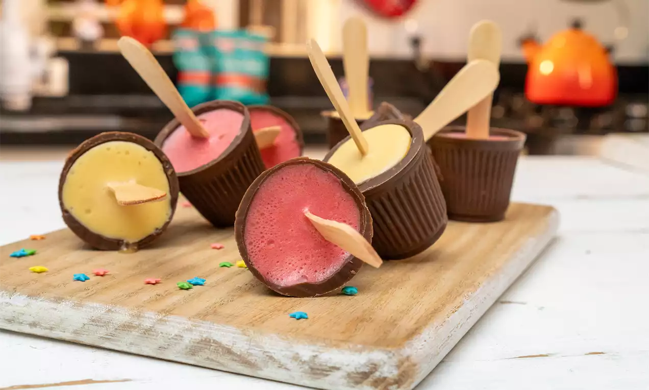 Copinhos de chocolate com gelatina cremosa amarela e rosa, decorados com palitos de madeira sobre tábua rústica e granulados coloridos