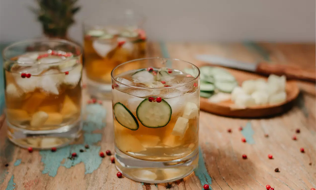 Chá gelado dourado com cubos de gelo, rodelas de pepino e grãos de pimenta rosa em copos baixos sobre mesa rústica com abacaxi ao fundo